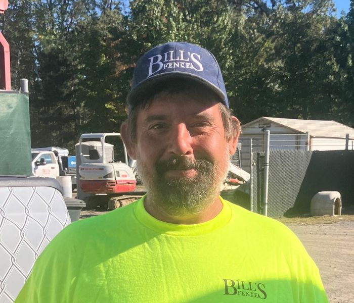 A man with a beard wearing a navy Bills Fences cap and a bright yellow Bills Fences shirt stands outdoors on a sunny day near machinery and storage buildings.