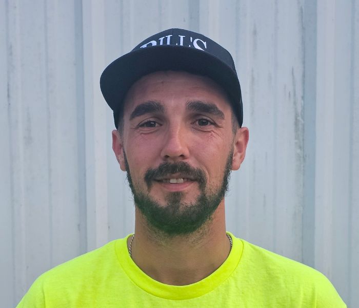 A man with a beard and mustache wearing a black cap and a bright yellow shirt stands in front of a corrugated metal wall, smiling gently at the camera.