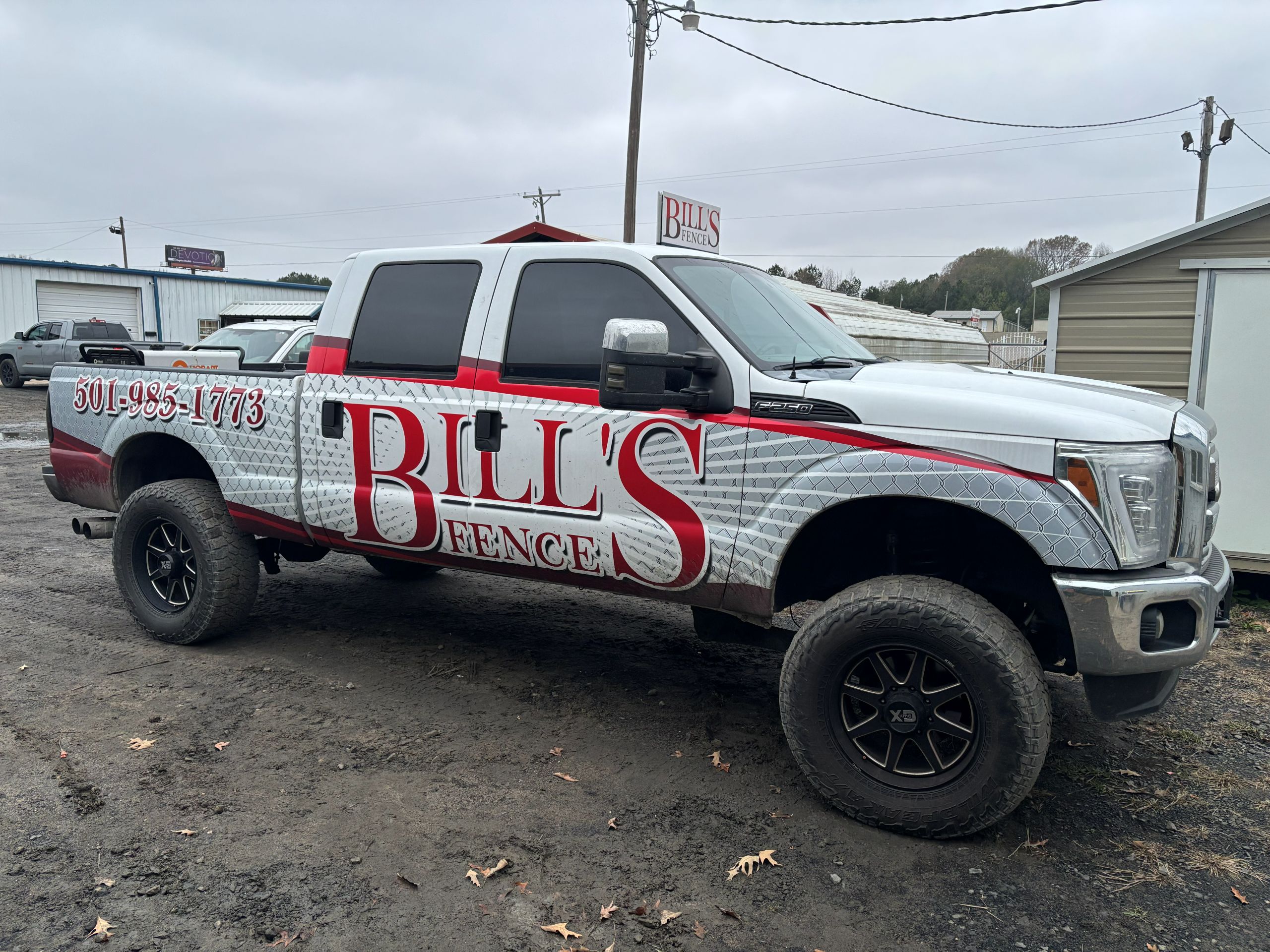 fence-company-warranty A silver pickup truck with Bills Fence and a phone number on the side is parked on a dirt lot near buildings and power lines under a cloudy sky.