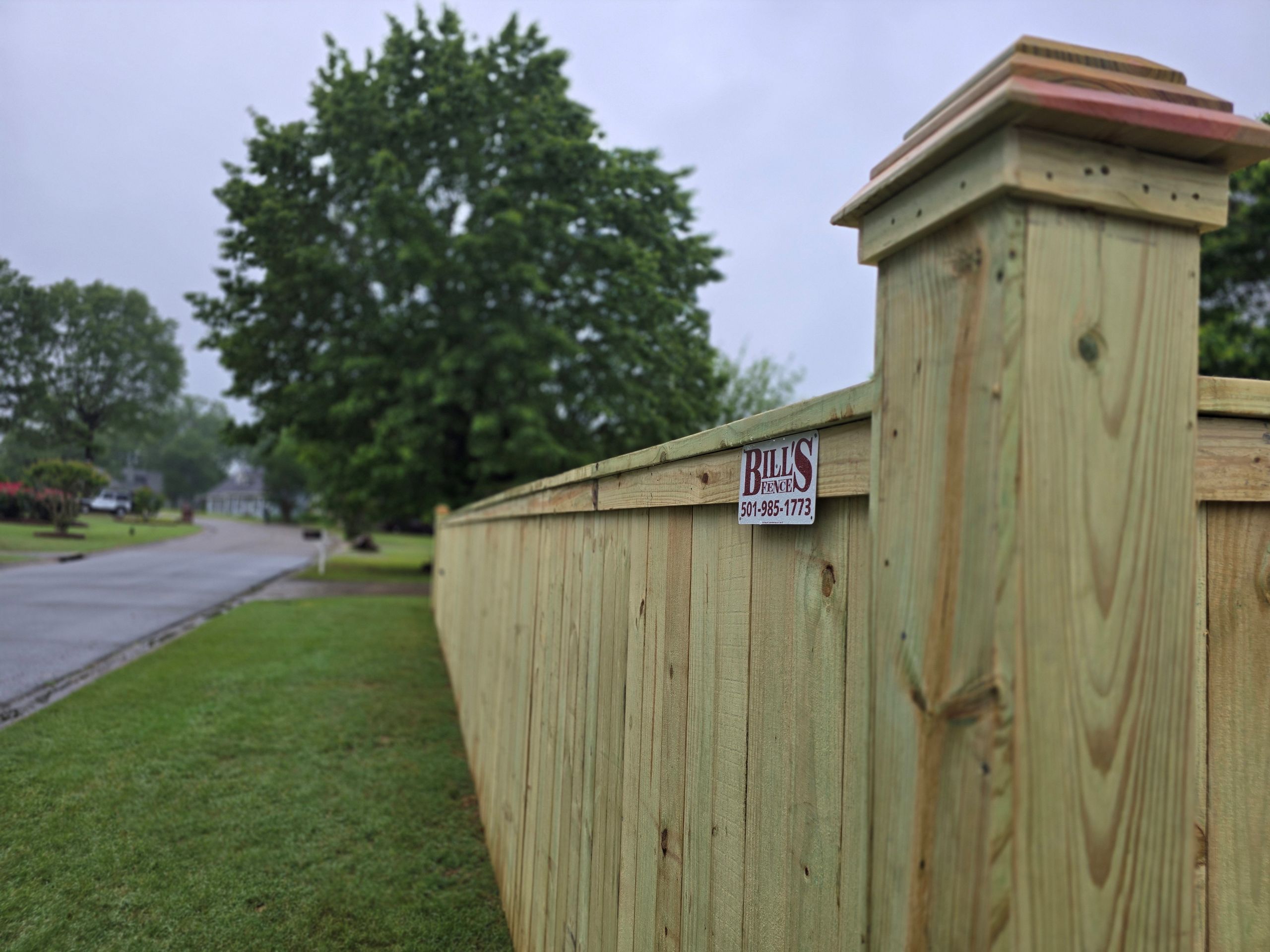 A newly built wooden fence runs alongside a quiet suburban street on a cloudy day, with a small sign reading Bills Fences attached to one post. Lush green grass and trees are visible in the background, enhancing curb appeal and potentially boosting property value in Arkansas.