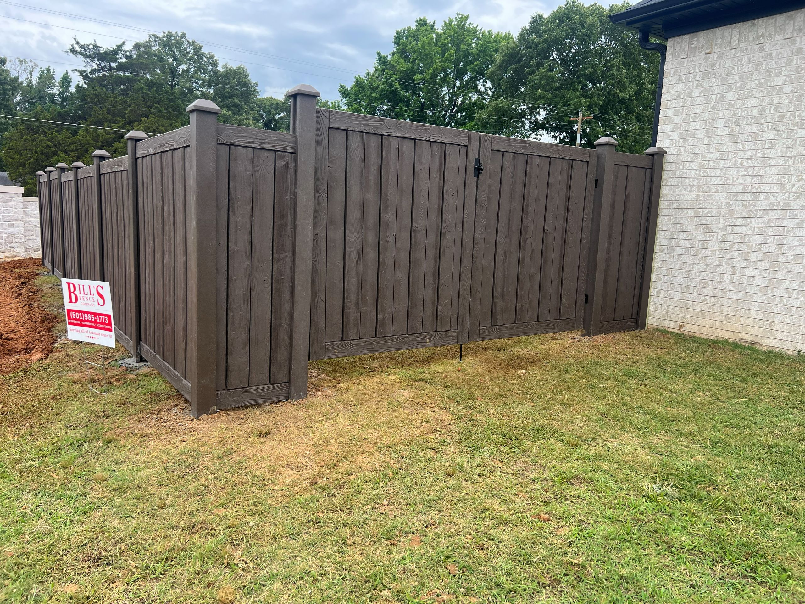 A brown wooden privacy fence enclosure stands beside a light brick building, with a real estate sign reading Bills in the grass nearby. Trees and a cloudy sky are visible in the background.