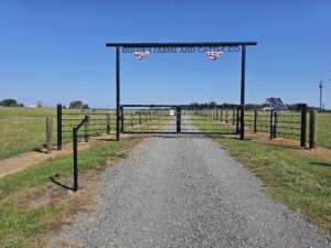 A gravel road leads through a metal gate with a sign reading HOLDEN FARMS AND CATTLE CO into a fenced grassy field under a clear blue sky. A solar panel is on the right side near the gate.