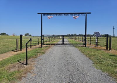 A gravel road leads through a metal gate with a sign reading HOLDEN FARMS AND CATTLE CO into a fenced grassy field under a clear blue sky. A solar panel is on the right side near the gate.