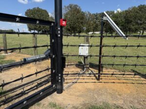 A solar-powered electric gate with barbed wire stands open on a rural dirt road, surrounded by grassy fields, trees, and a blue sky. A small white control box and solar panel are mounted nearby.