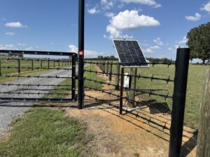 A metal gate with barbed wire and a solar panel stands at the entrance of a fenced rural road, surrounded by green grass and fields under a blue sky with scattered clouds.