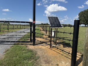 A black metal gate with electric wires and a solar panel by a rural gravel road, surrounded by green grass and open fields under a blue sky with clouds.