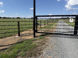 A metal gate with star decorations blocks a gravel driveway leading to a grassy field, fenced with wooden posts and wire, under a blue sky with scattered clouds.