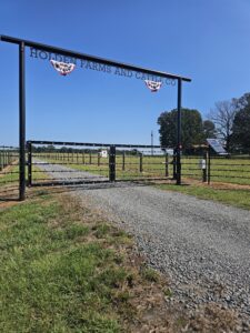 A gravel driveway leads to a gated entrance reading HOLDEN FARMS AND CATTLE CO. The gate is decorated with patriotic bunting and surrounded by green pastures and trees under a clear blue sky.