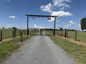 A gravel driveway leads through a metal gate labeled Holden Farms and Cattle Co. Fields of green grass lie on both sides under a blue sky with scattered clouds.