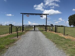 Gravel driveway leads to a fenced entrance with a sign reading Holden Farms and Cattle Co over a metal gate; open fields and a few trees are visible under a blue sky with scattered clouds.