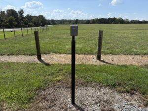 A keypad stands on a black post in front of an open grassy field, with two wooden posts and a dirt path behind it under a mostly sunny sky.