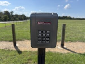 A LiftMaster keypad is mounted on a post in front of a grassy field, with a road and trees visible in the background under a blue sky with clouds.