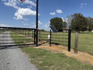 A black metal gate with a solar panel and keypad blocks a gravel driveway leading to a house; grassy fields and trees are visible under a blue sky with scattered clouds.