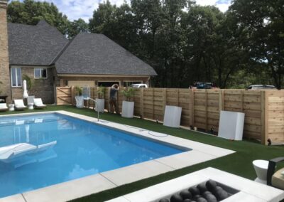 A backyard with a swimming pool, white lounge chairs, a wooden fence, and some scattered panels. A person stands near the fence, and trees fill the background.