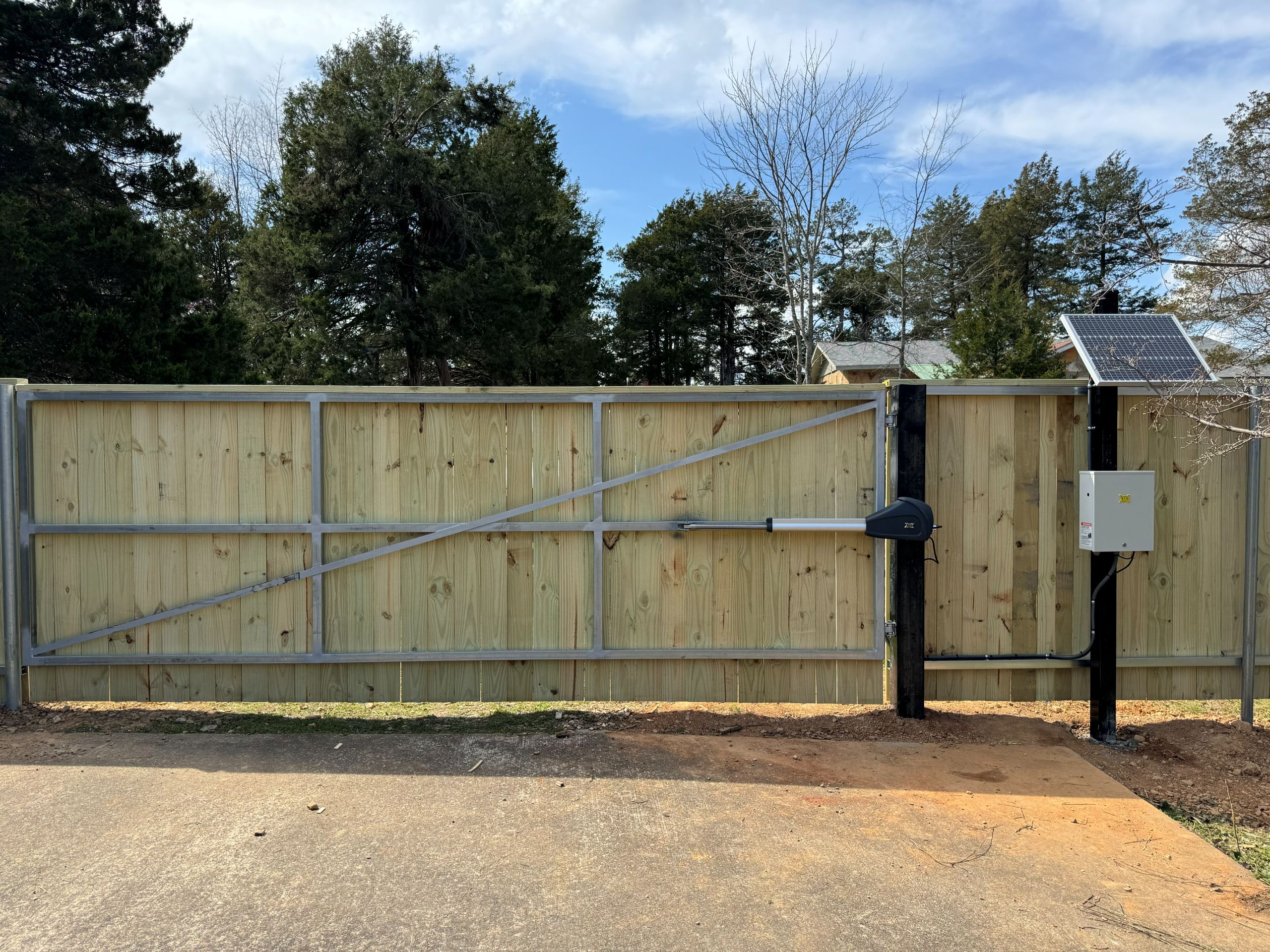 A wooden privacy gate with a metal frame and automatic opener, mounted between black posts. A solar panel and control box are attached to the right post, with trees and a house visible in the background.
