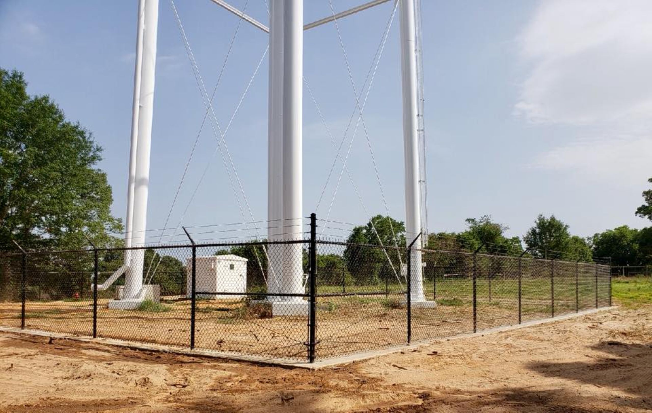 A tall white water tower is surrounded by permanent fencing and dirt ground, with some trees and a small white utility building in the background under a partly cloudy sky.