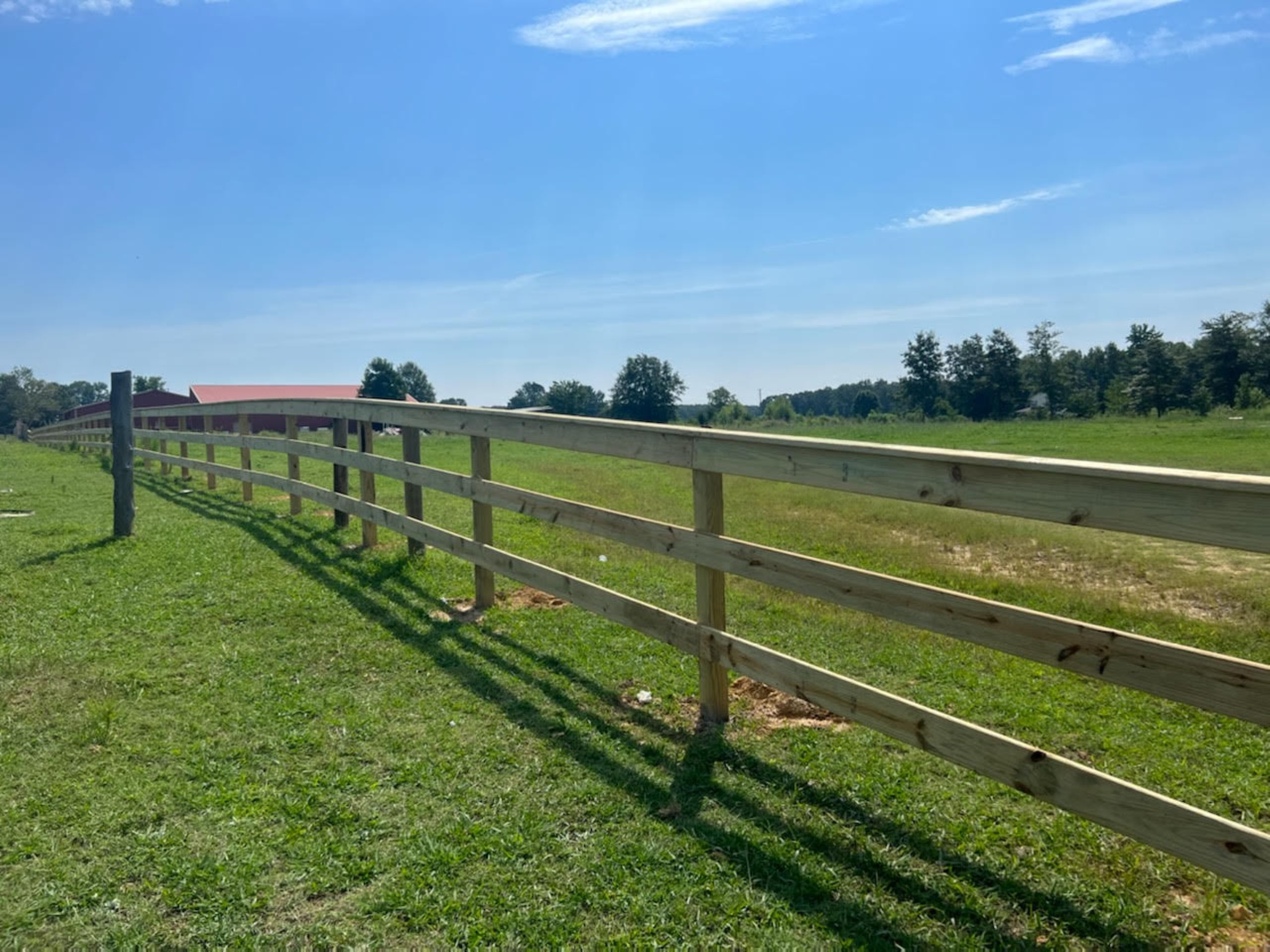 A wooden three-rail fence curves across a grassy field toward a red barn under a clear blue sky.