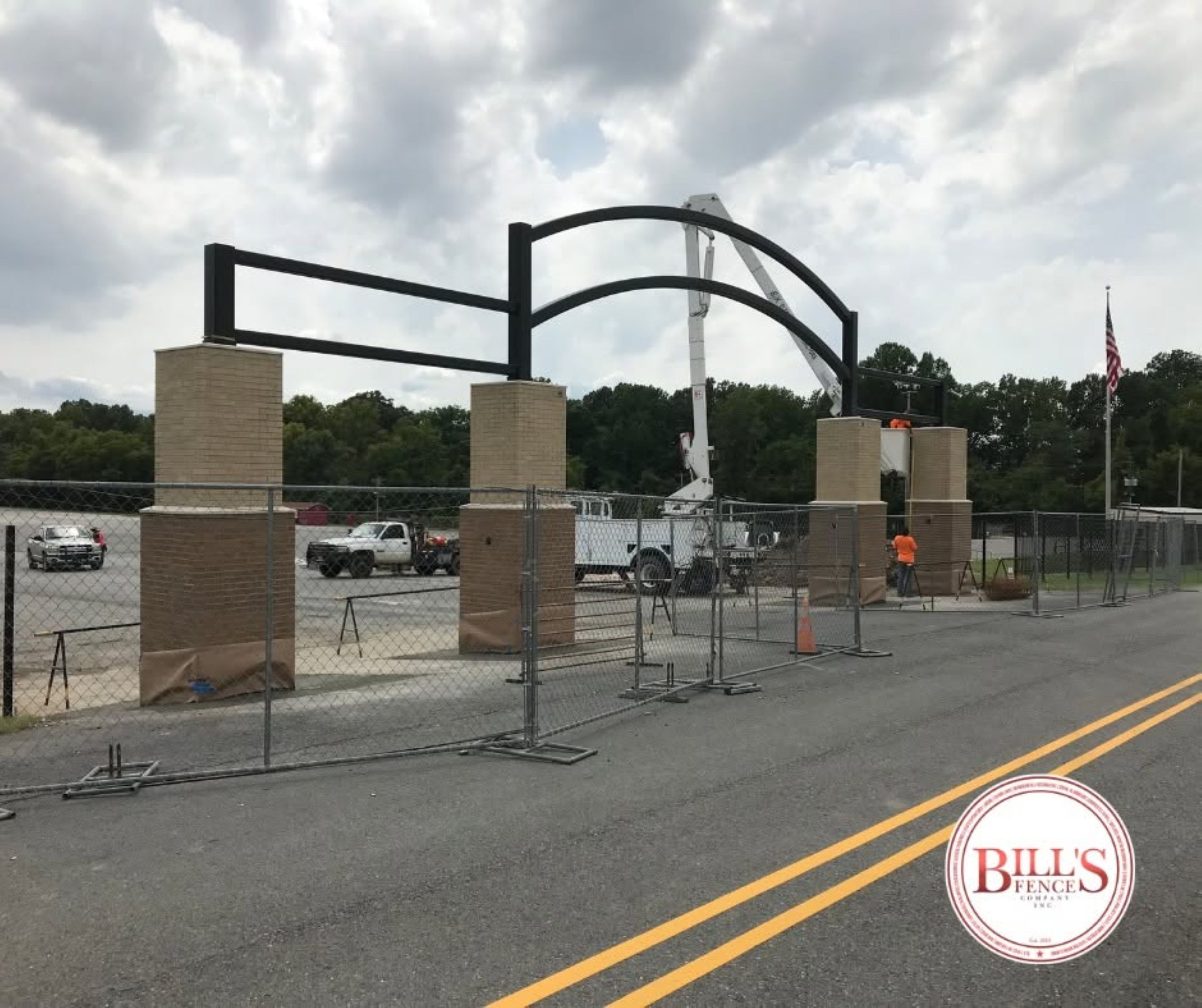 Construction crew using a boom lift to install a large black metal archway between brick columns at a site enclosed by temporary chain-link fencing.